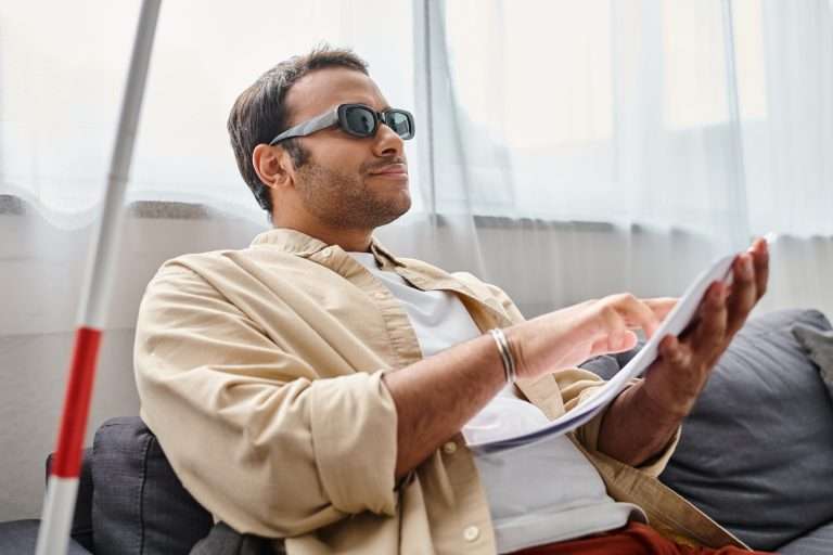 cheerful indian man with visual impairment with glasses sitting o sofa and reading braille code